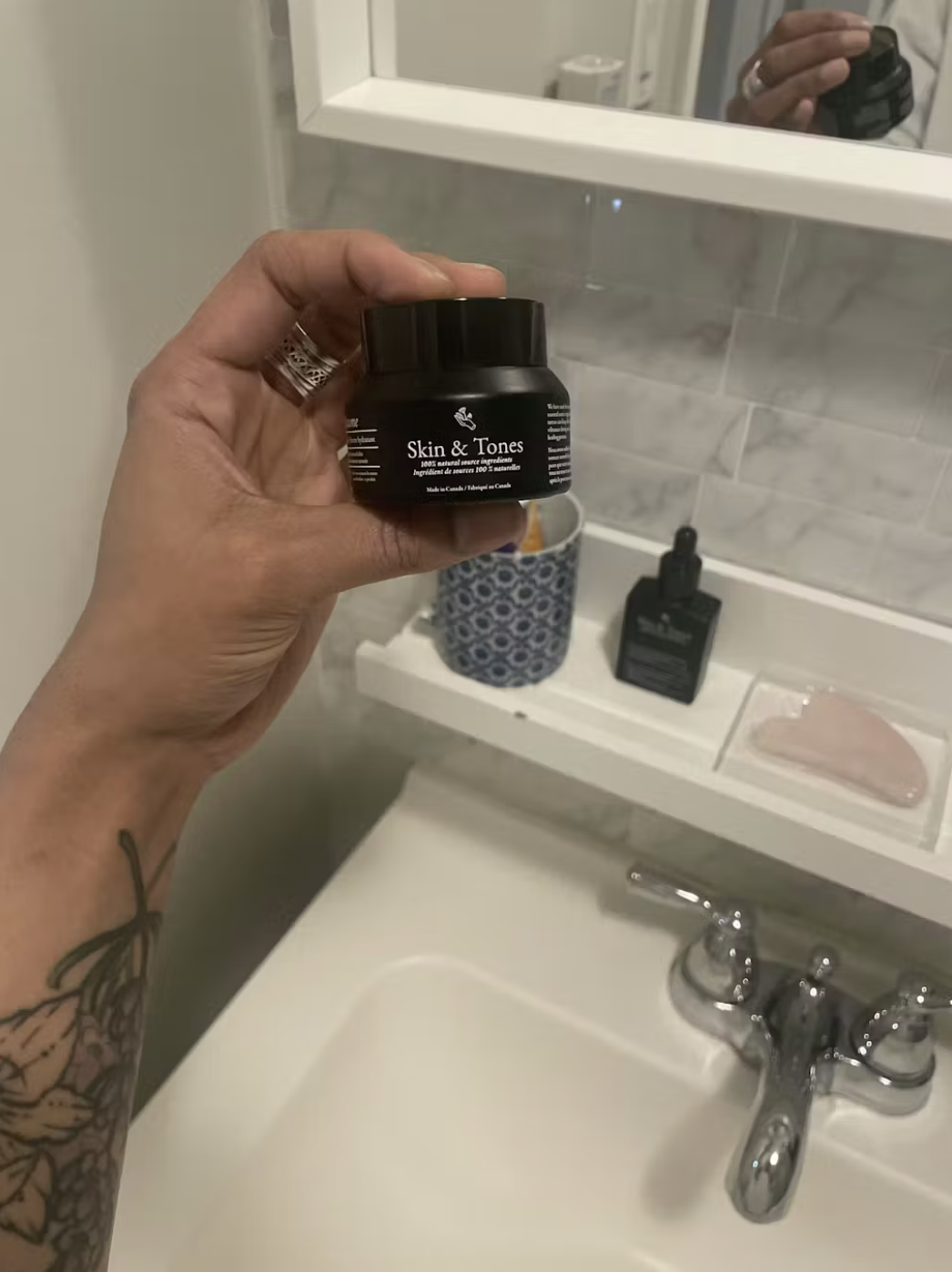 A person with a tattooed arm holds up a small black jar of Skin & Tones cream.