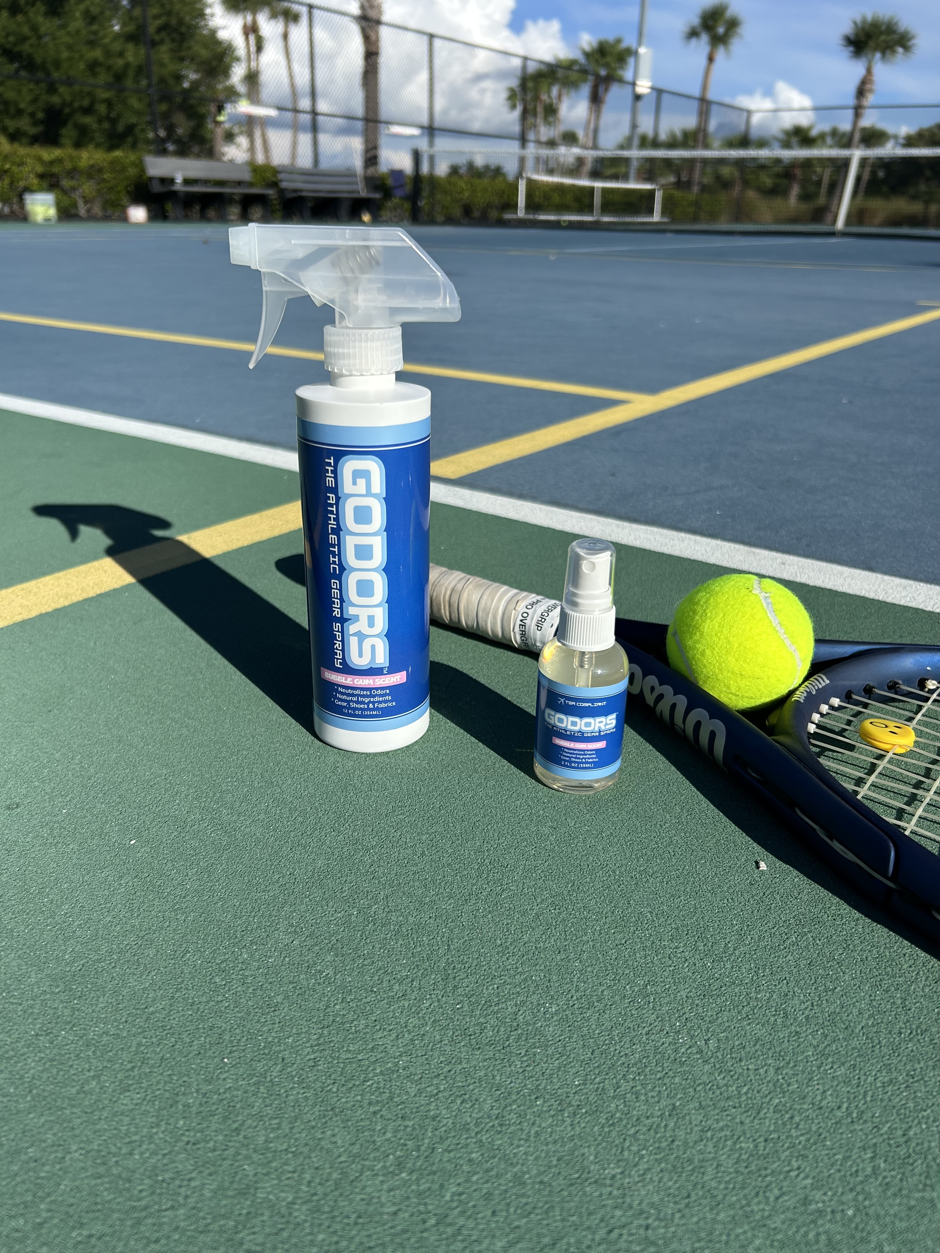 Godors athletic gear spray bottles, a tennis racquet, and a ball on an outdoor tennis court.