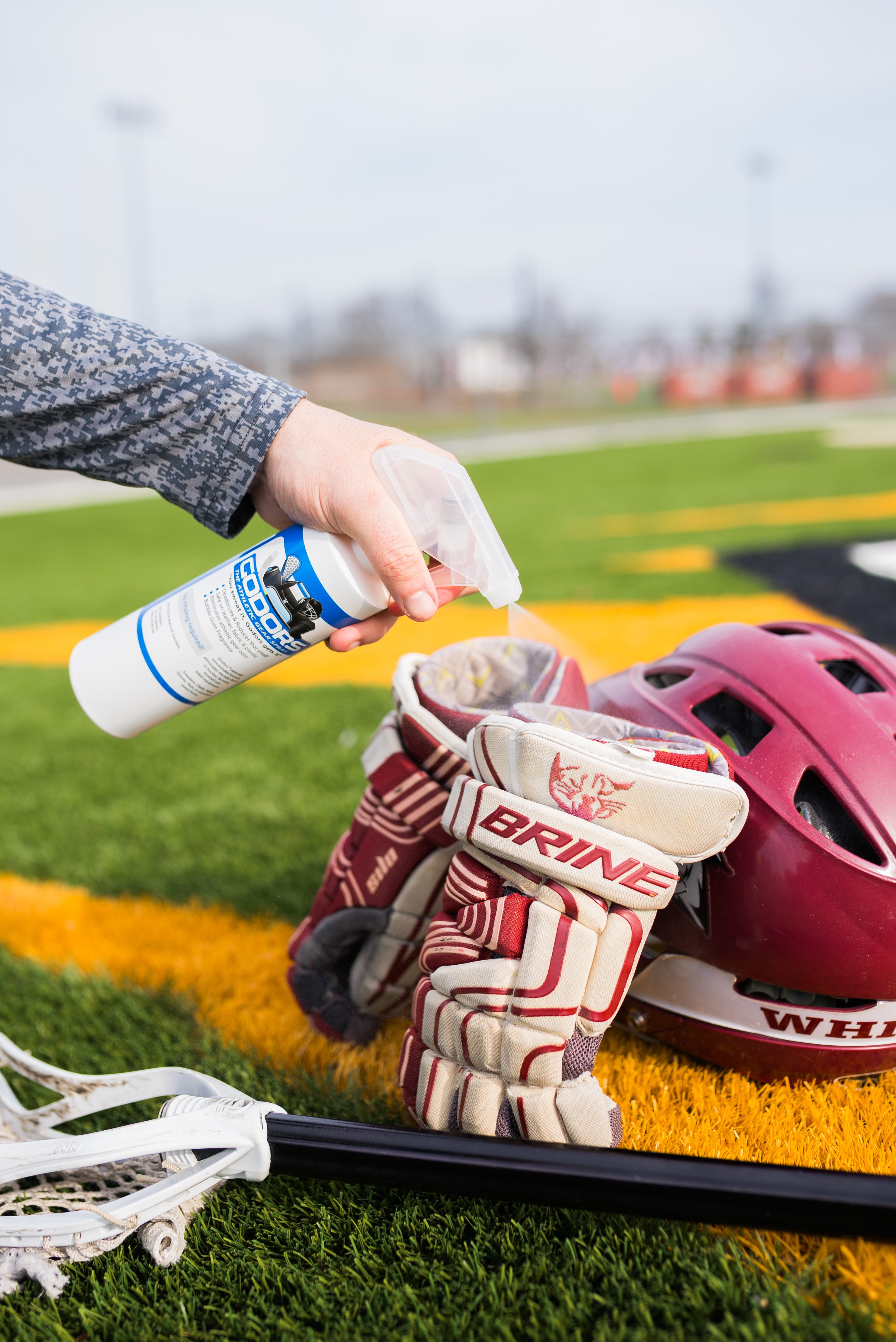 A person sprays lacrosse gloves on a turf field next to a helmet and stick.