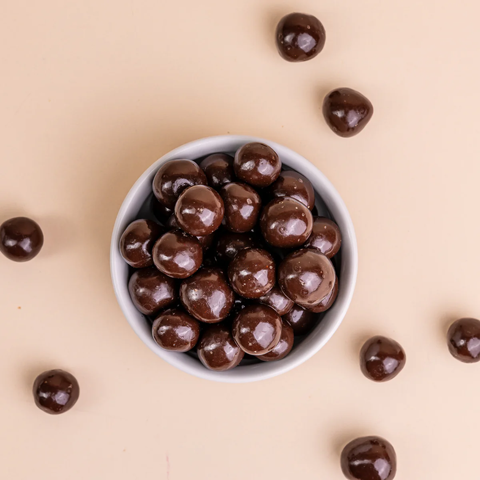 An overhead view of a small bowl of shiny, dark chocolate balls on a light beige background.