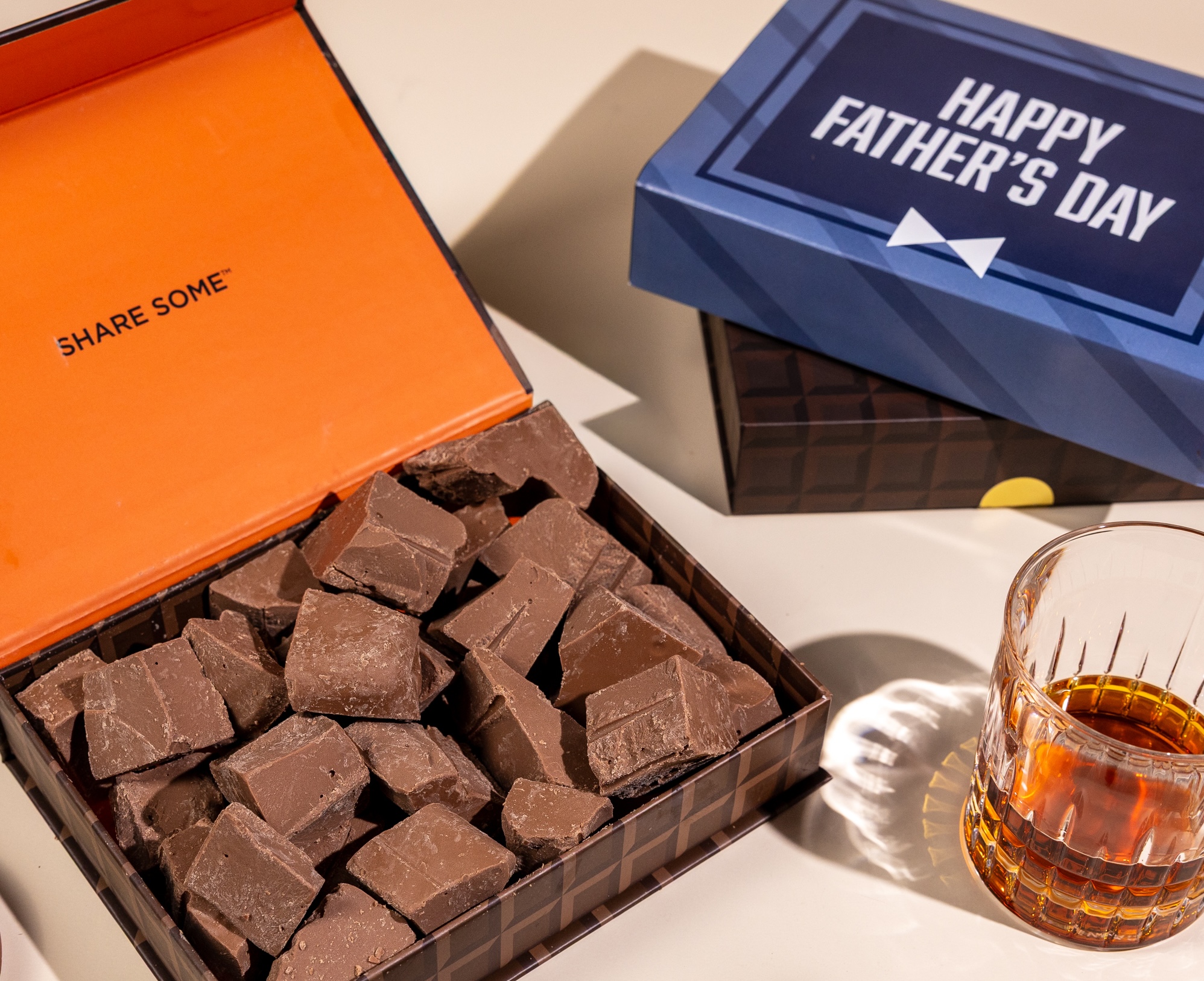 An open box of chocolate chunks sits next to a glass of whiskey and a Father's Day gift box.