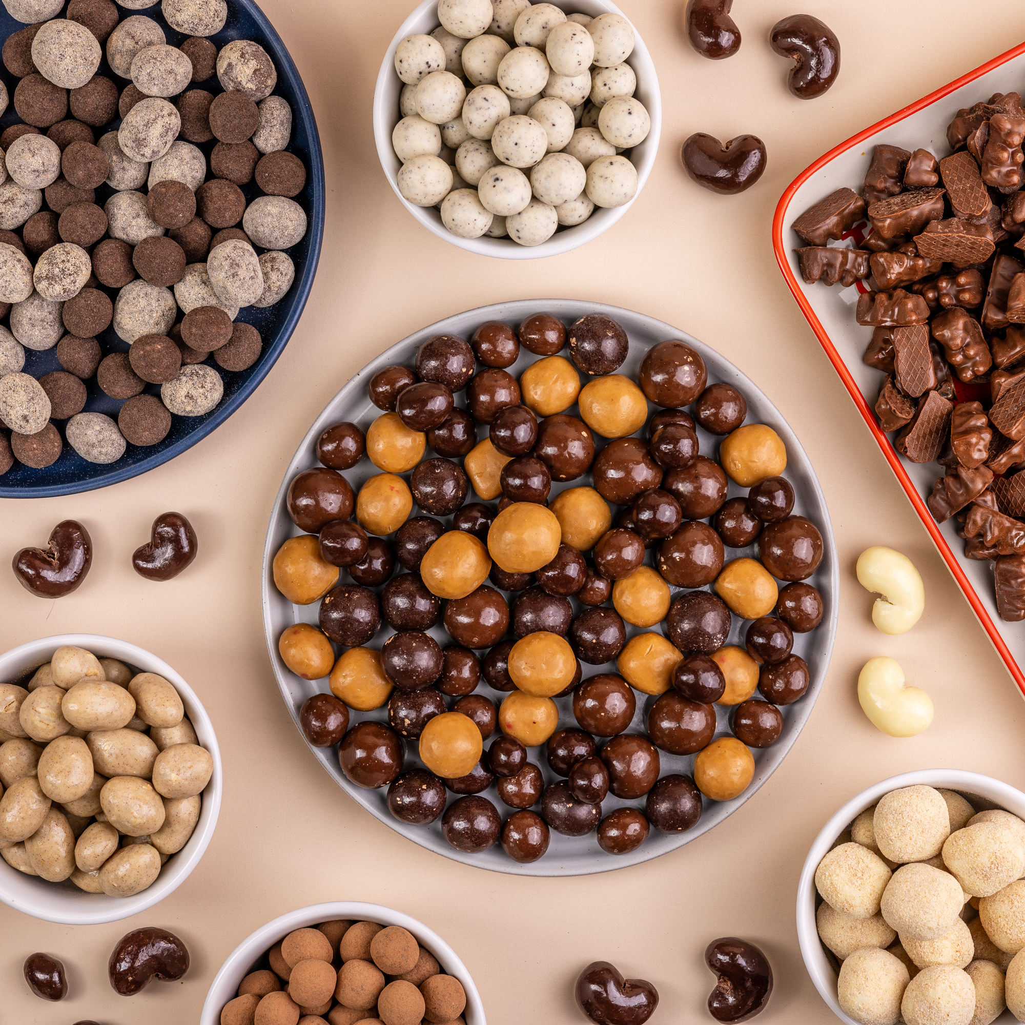 Assorted chocolate-covered nuts and candies in bowls and trays on a beige background.