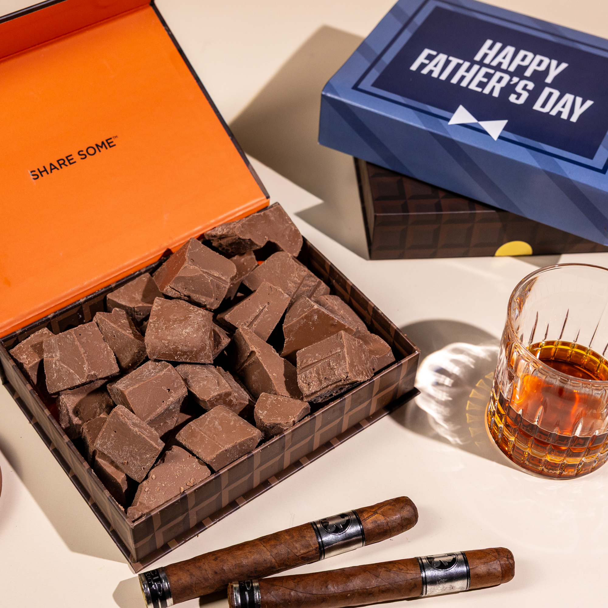 A Father's Day gift set with a box of chocolate chunks, two cigars, and a glass of liquor.
