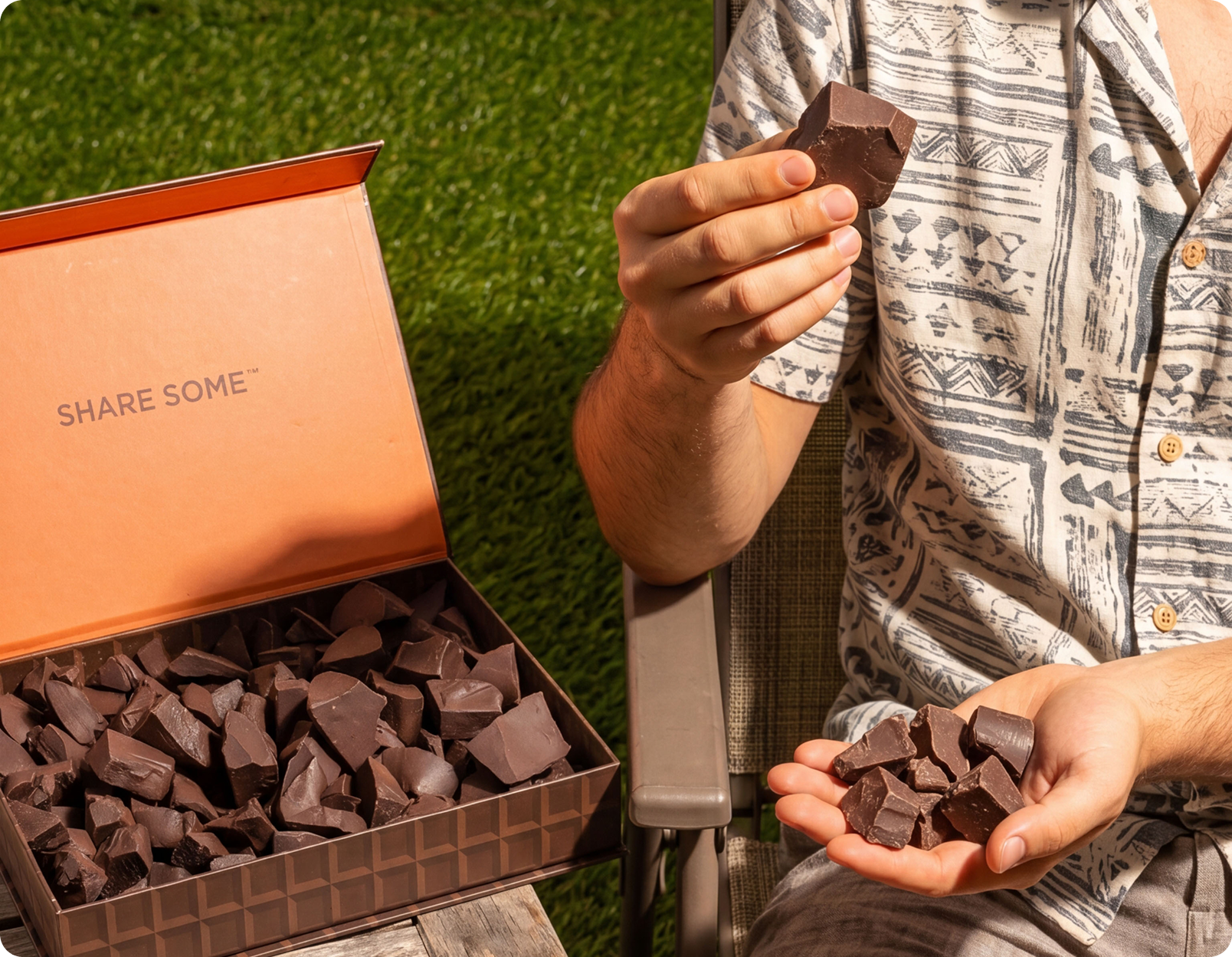A person sitting outside holds chunks of dark chocolate from a large box full of chocolate pieces.