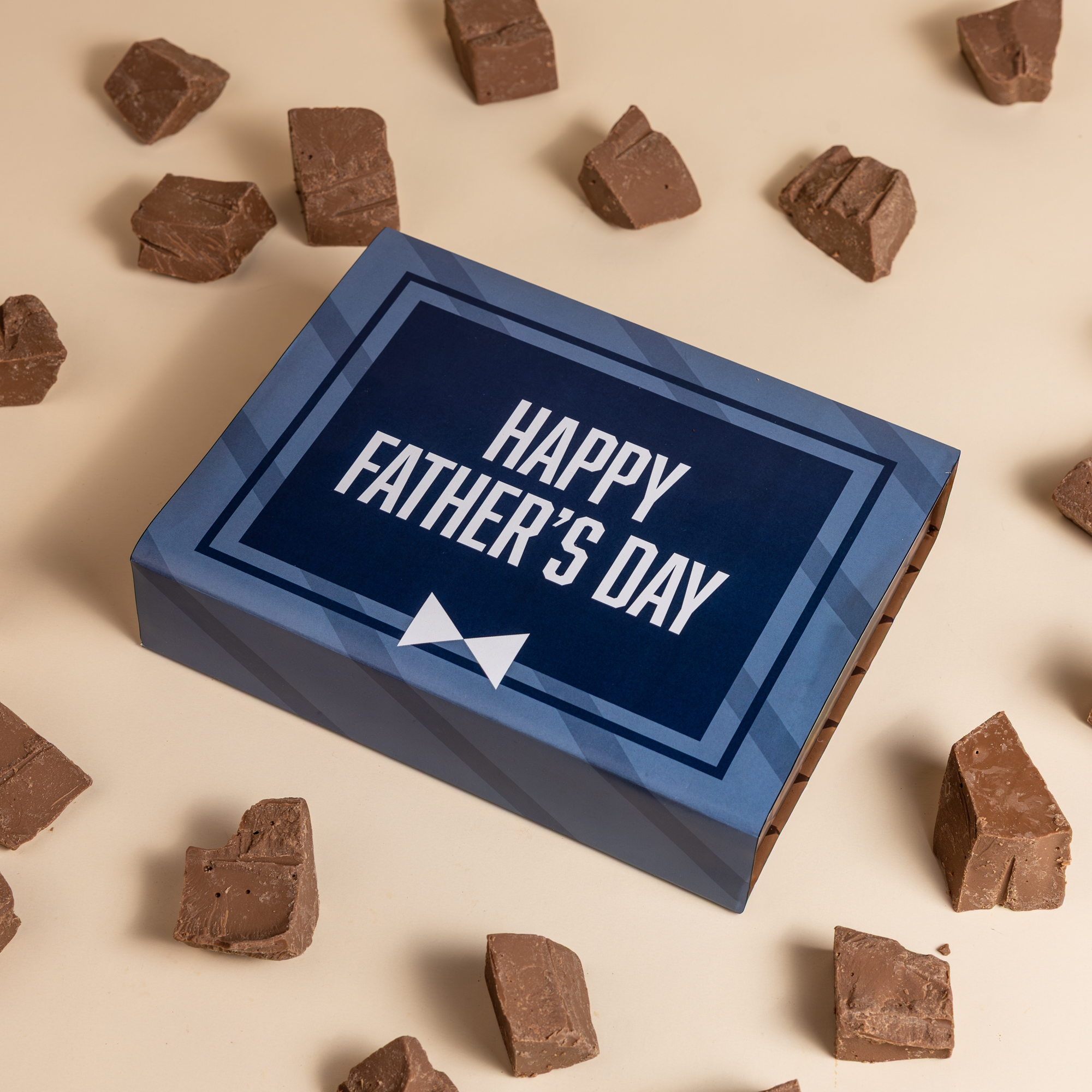 A blue 'Happy Father's Day' gift box is surrounded by chunks of chocolate fudge on a beige surface.