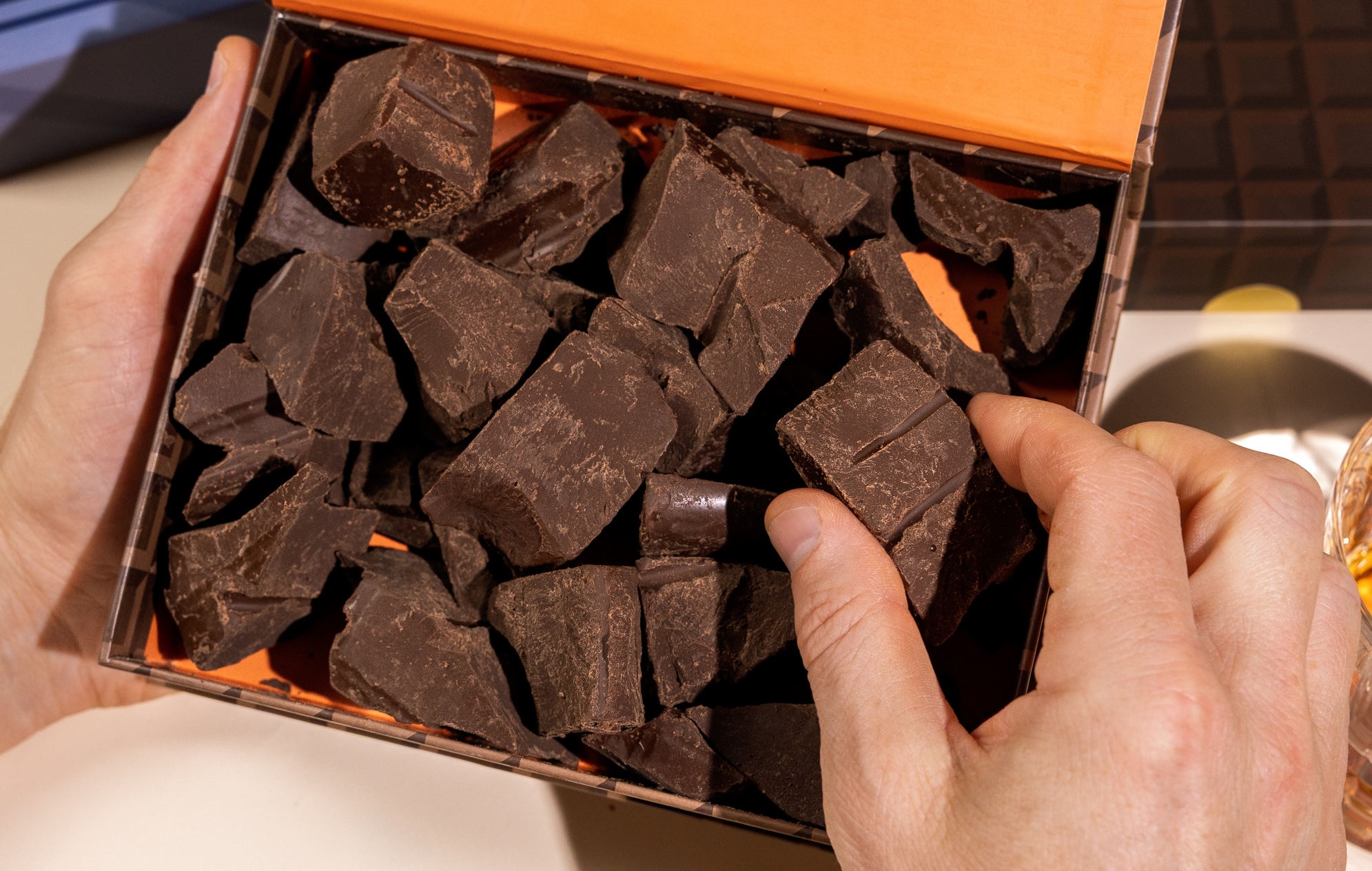 A person's hands hold an open box filled with large, rough chunks of dark chocolate.