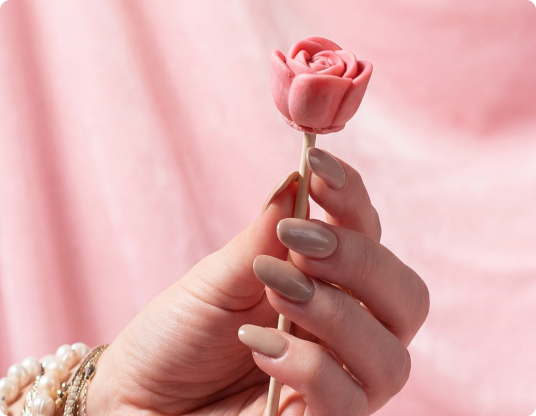 A hand with beige manicured nails holds a pink rose-shaped candy against a pink draped background.