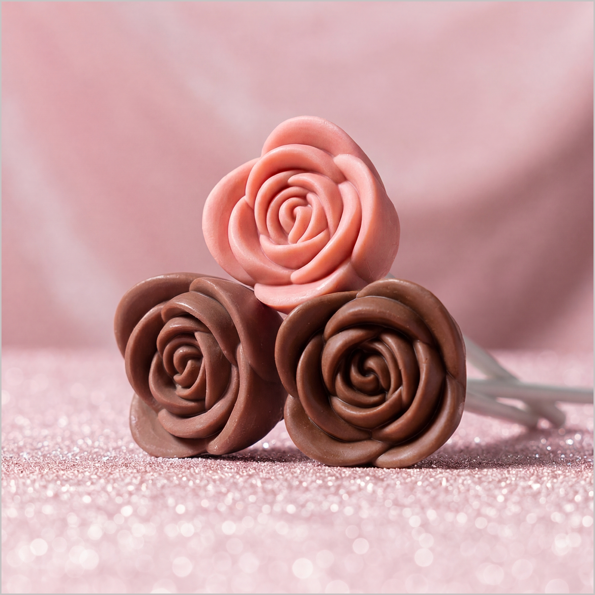Three rose-shaped chocolate lollipops, one pink and two brown, on a sparkly pink surface.