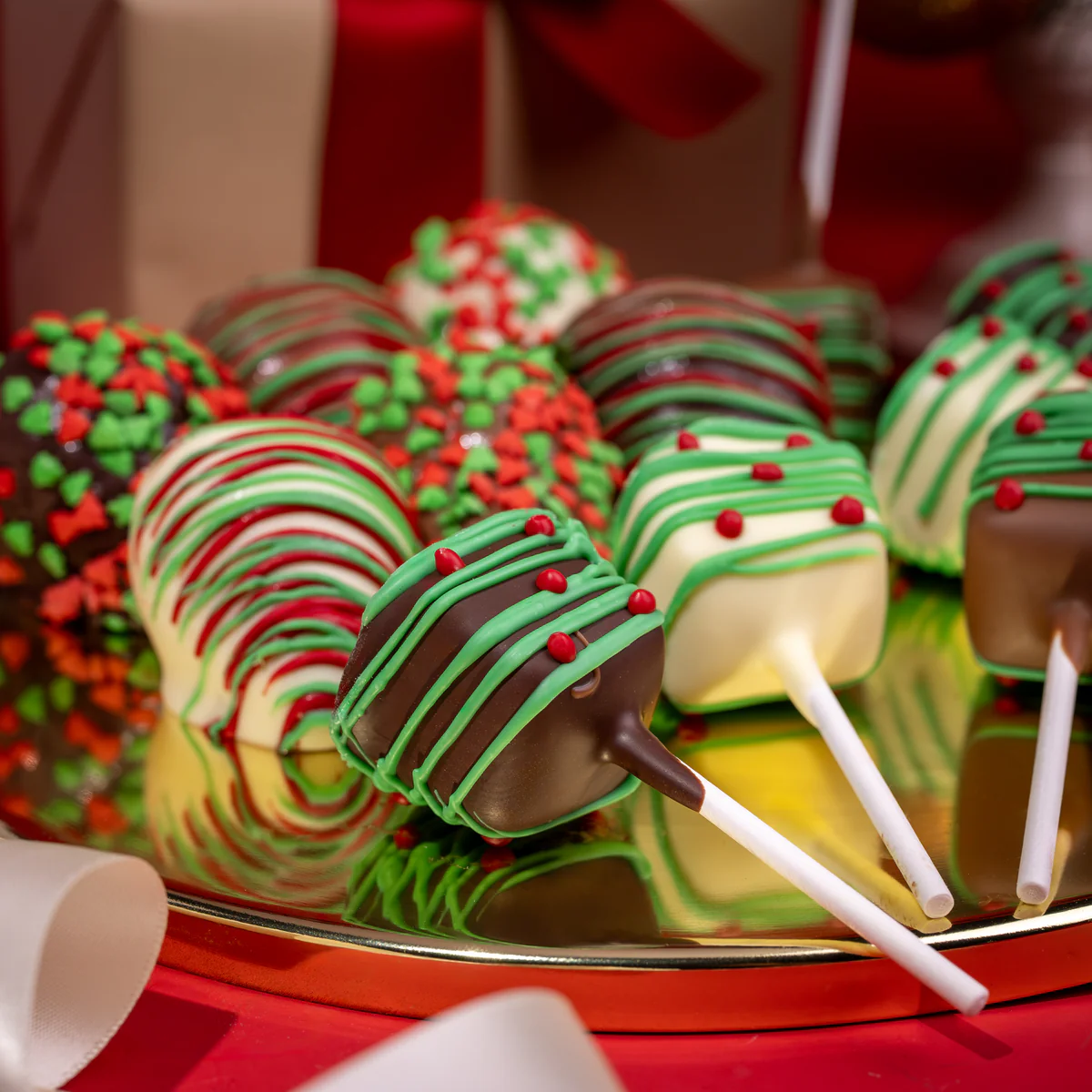 Festive cake pops with colorful icing and sprinkles on a reflective plate.