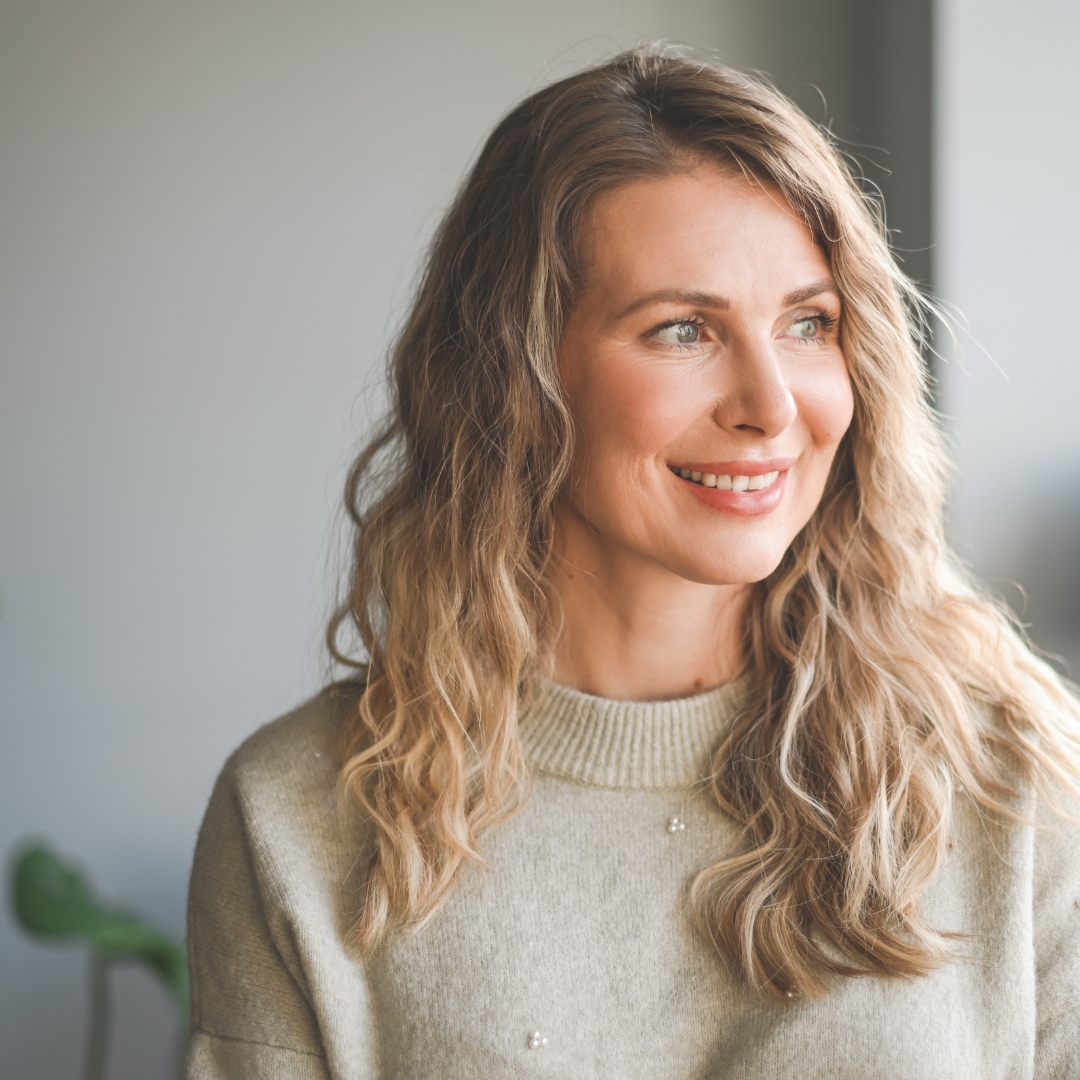 Smiling person with long wavy hair wearing a light sweater, looking to the side.