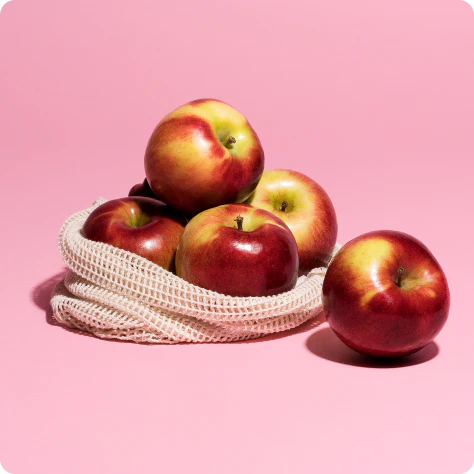 Several red and yellow apples sit in a white mesh bag on a pink background.