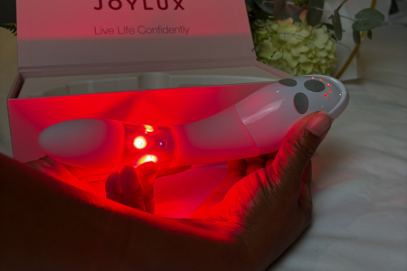A hand holding a white device emitting red light, with a box labeled 'Joylux' in the background.