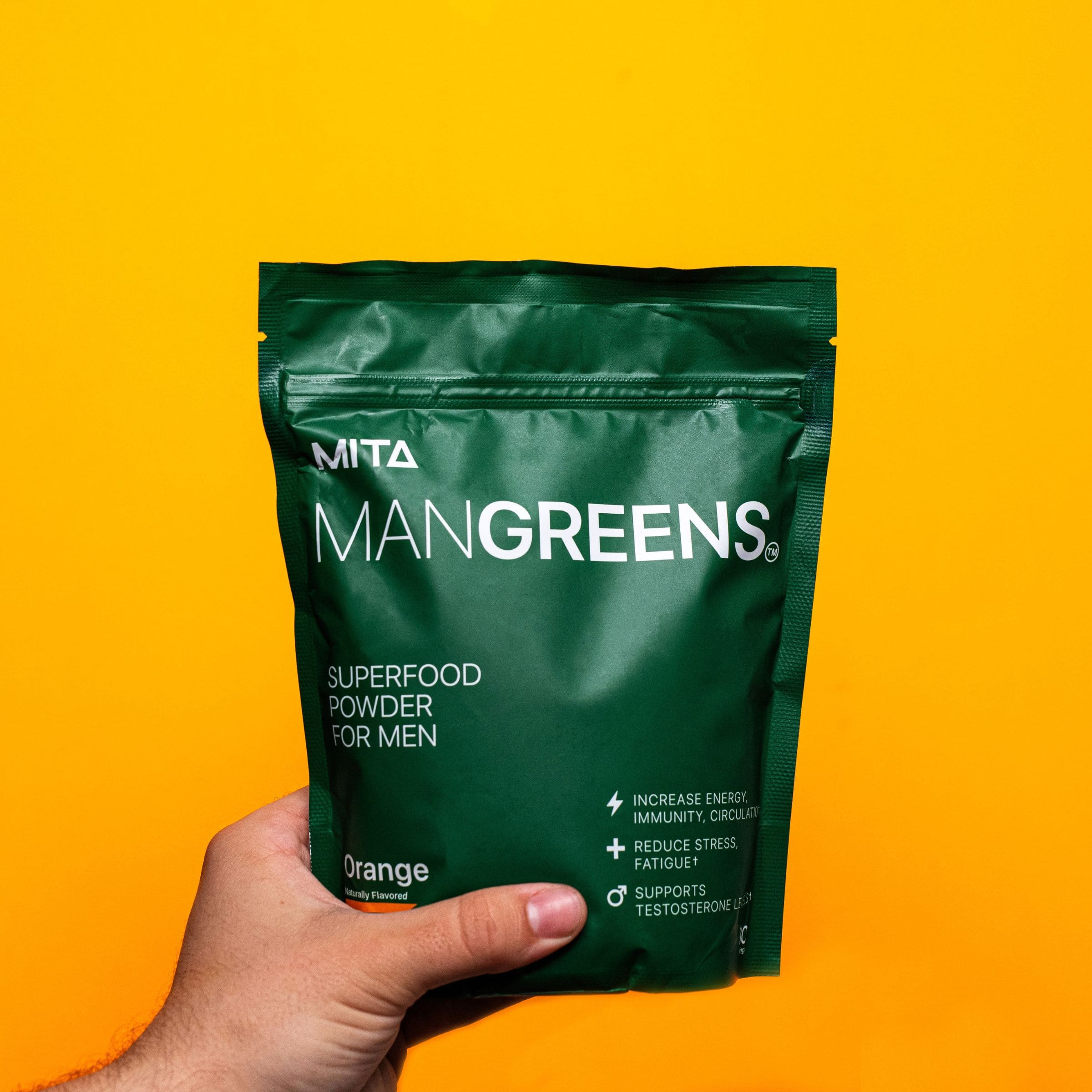 Hand holding a green bag of ManGreens superfood powder against yellow background.
