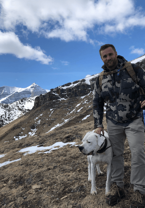 Man standing with a white dog on a mountain trail, with snowy peaks in the background.