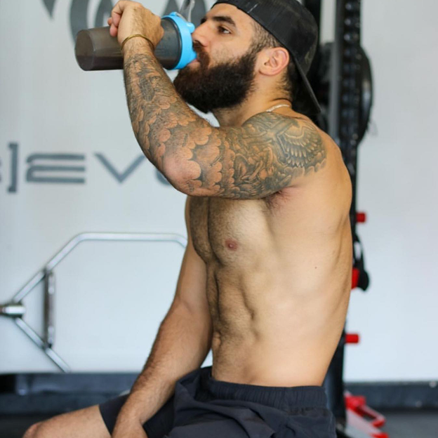 Man with tattoos drinking from a shaker bottle in a gym.
