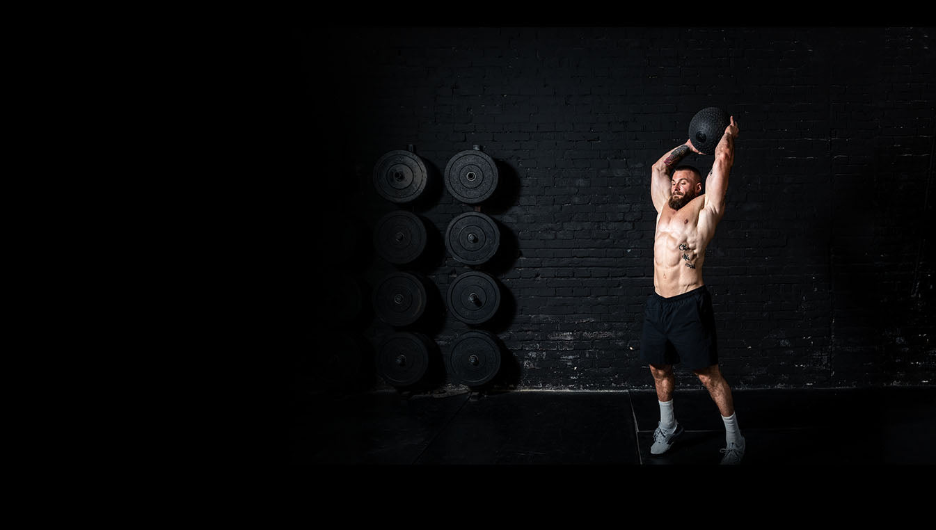 Man lifting a medicine ball in a gym with weights on the wall.