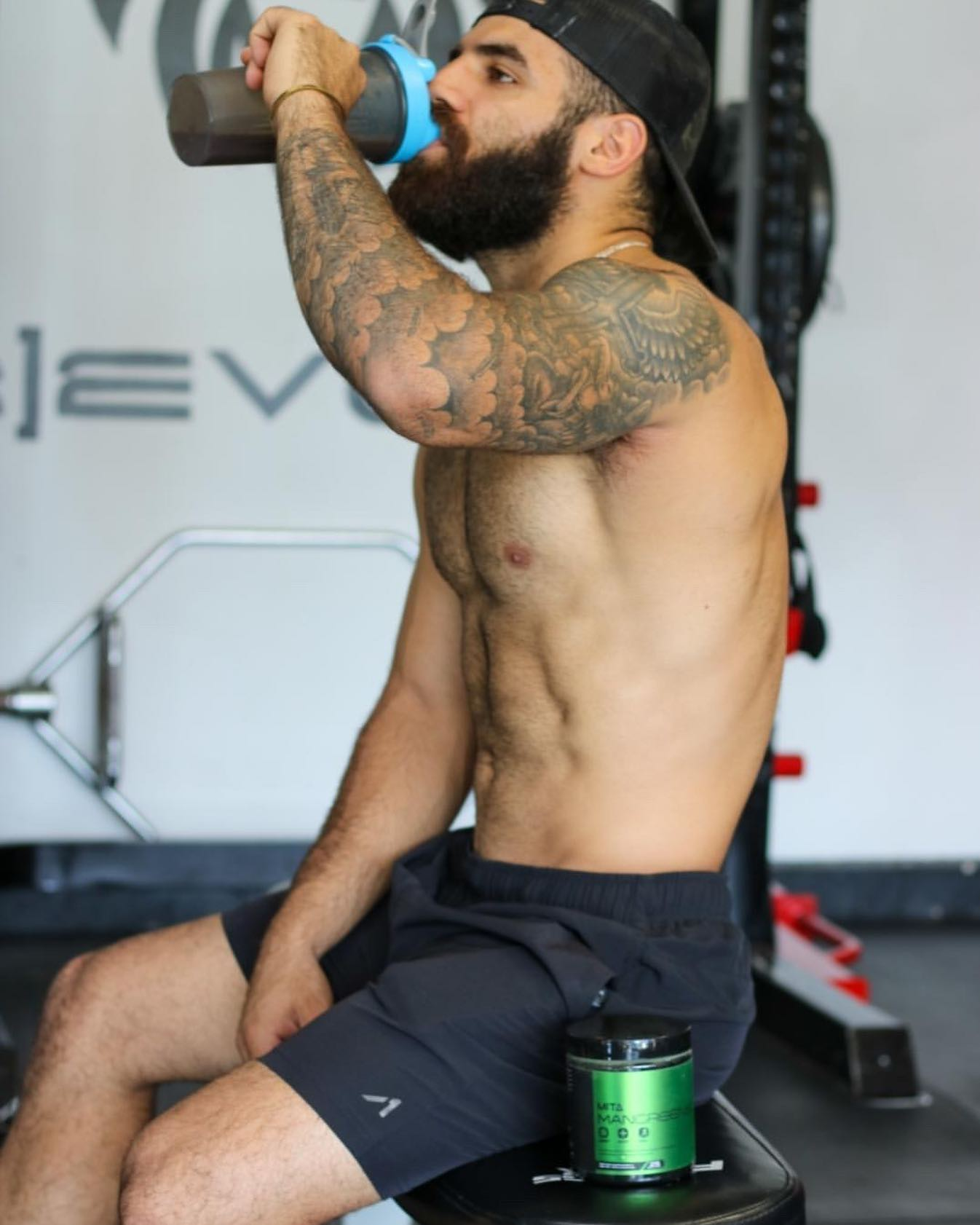Man with tattoos drinking from a shaker bottle in a gym setting.