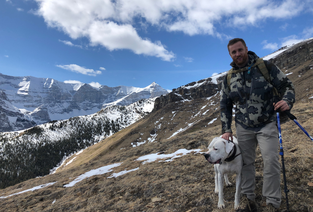 A man and a white dog hiking on a mountainside with snow-covered mountains in the background.