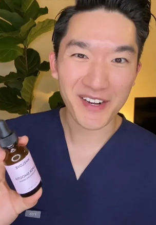 Person holding a skincare product bottle, smiling, with a plant in the background.
