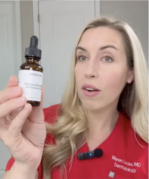 A woman in red scrubs holds up a small brown bottle of skincare serum with a white label.
