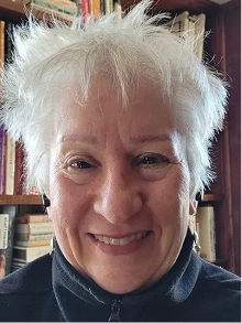 A smiling person with short, spiky white hair stands in front of a bookshelf.