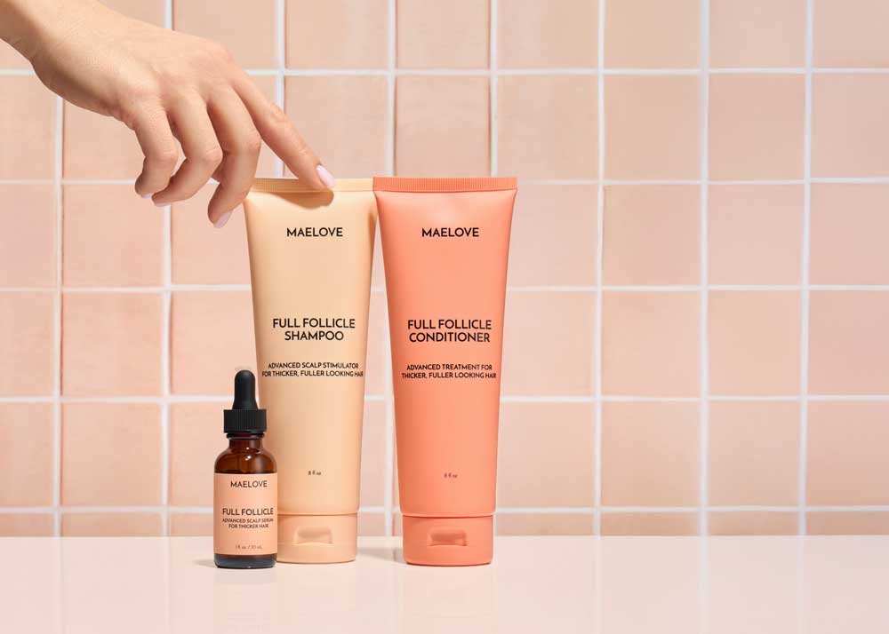 A hand reaches for Maelove's Full Follicle shampoo, conditioner, and scalp serum against a pink tiled wall.