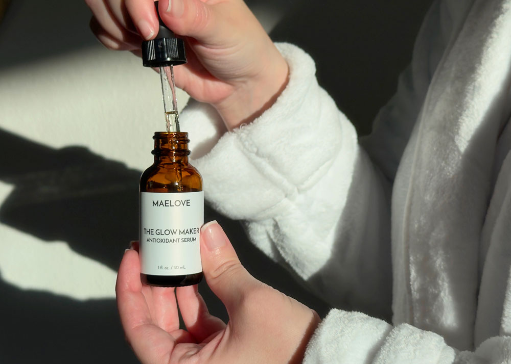A person in a white robe holds a bottle of Maelove The Glow Maker serum, using the dropper.