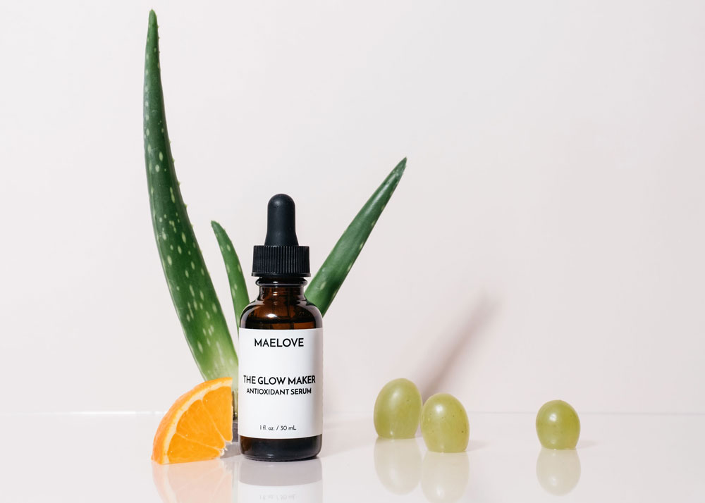 A bottle of Maelove antioxidant serum with aloe vera leaves, an orange slice, and green grapes.