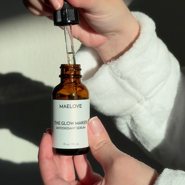 A person in a white robe holds a bottle of Maelove The Glow Maker serum with its dropper.