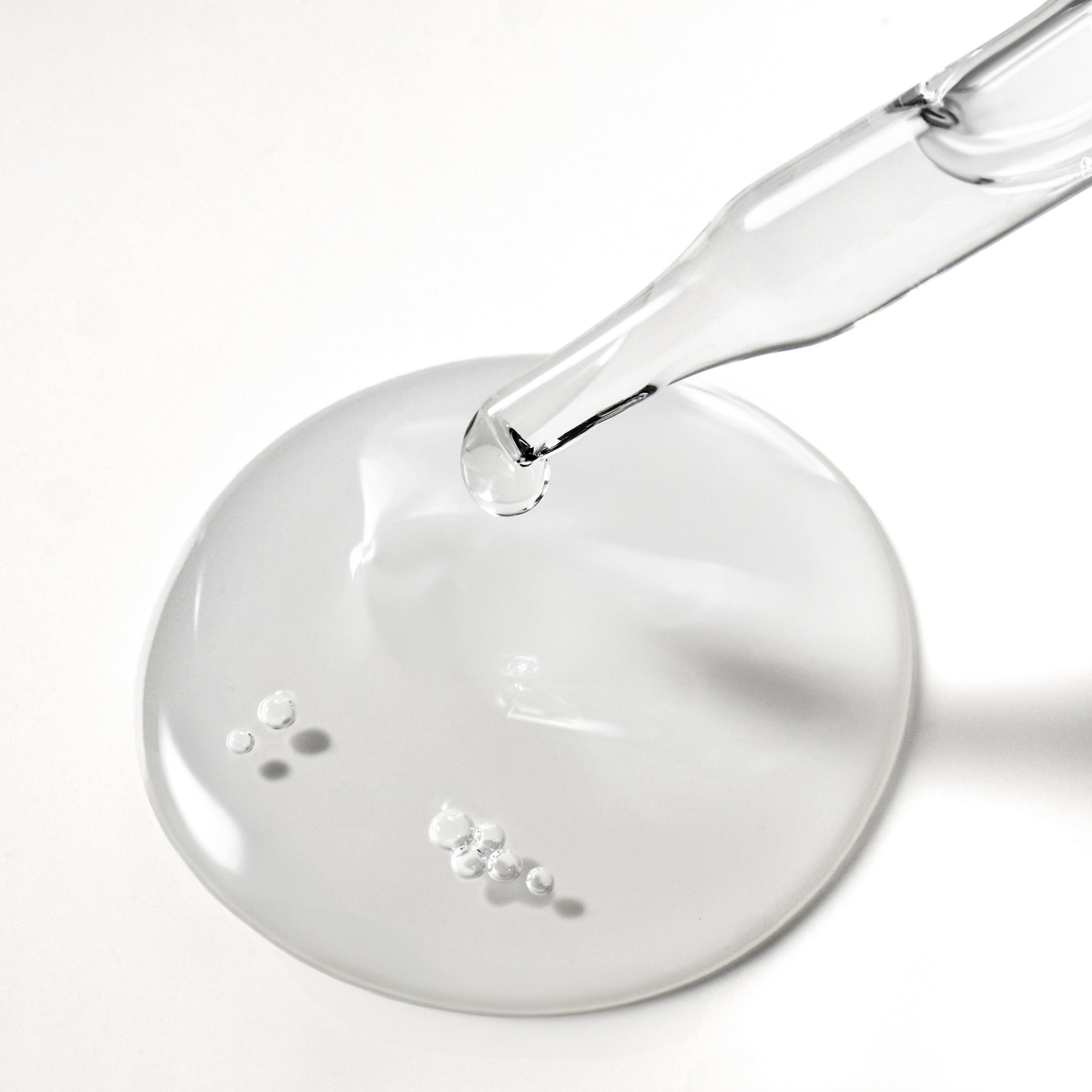 A close-up of a glass dropper dispensing clear liquid into a puddle on a white surface.