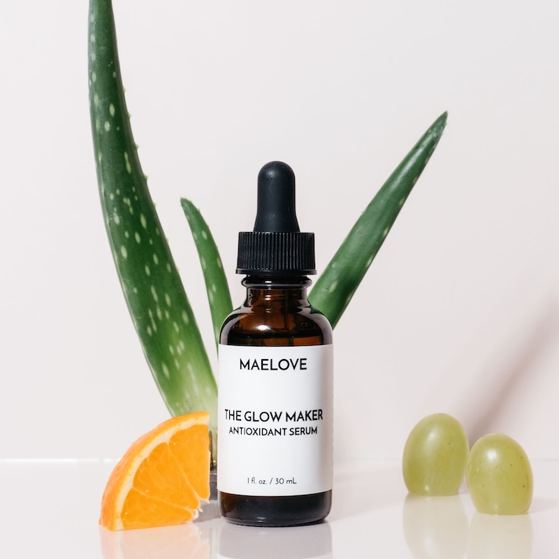 A bottle of Maelove The Glow Maker serum with aloe, an orange slice, and grapes on a white surface.