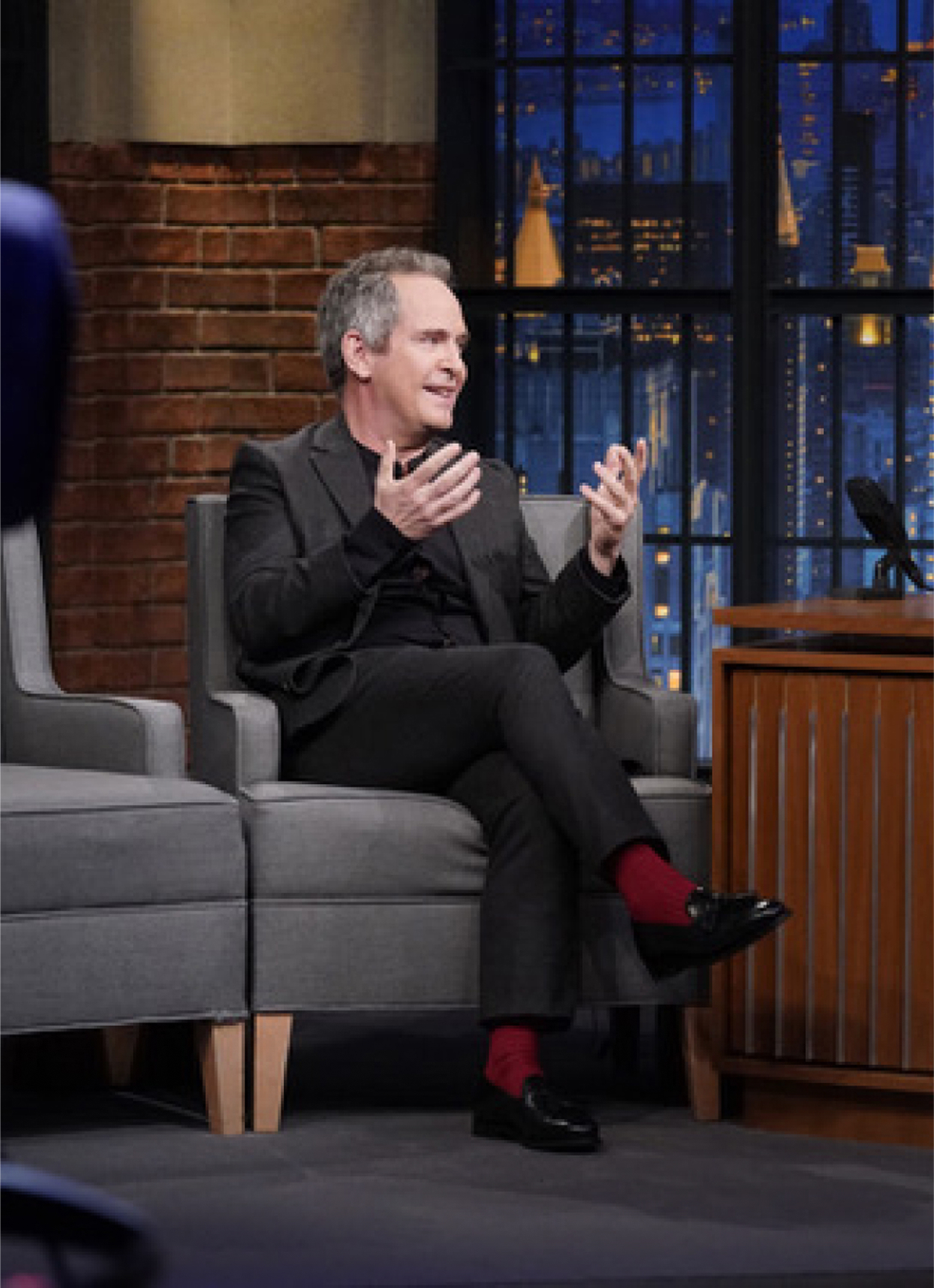 A man in a suit speaking on a nighttime talk show set.