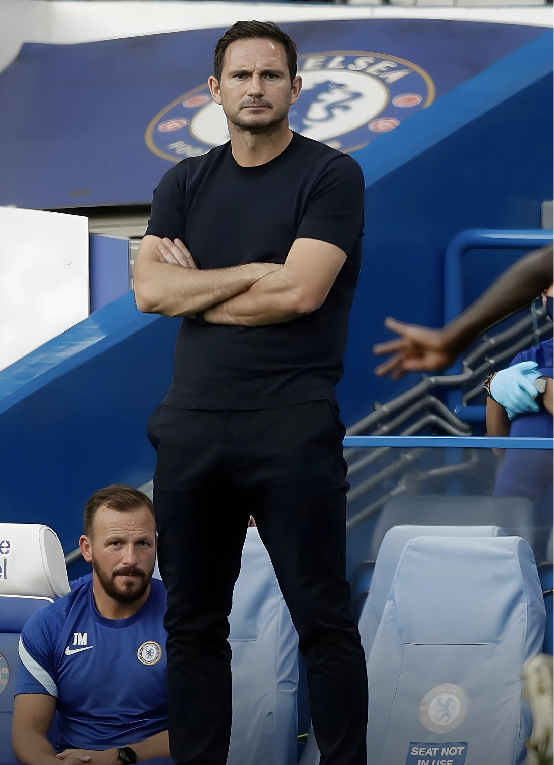 A man in black stands, arms crossed, on a sports sideline.