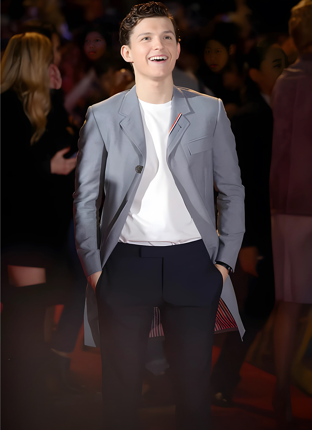 A smiling person in a grey coat on a red carpet event.