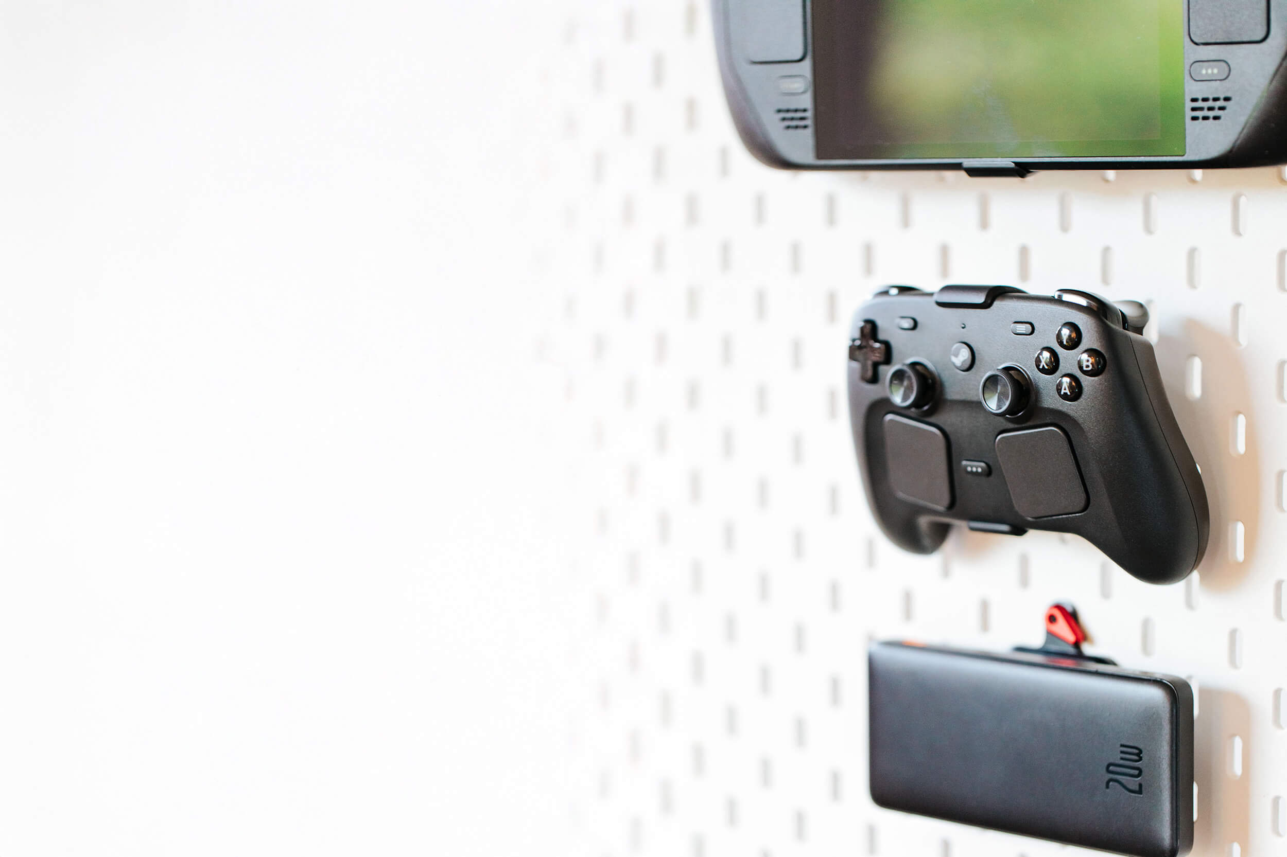 A handheld gaming console, a controller, and a power bank mounted on a white pegboard wall.