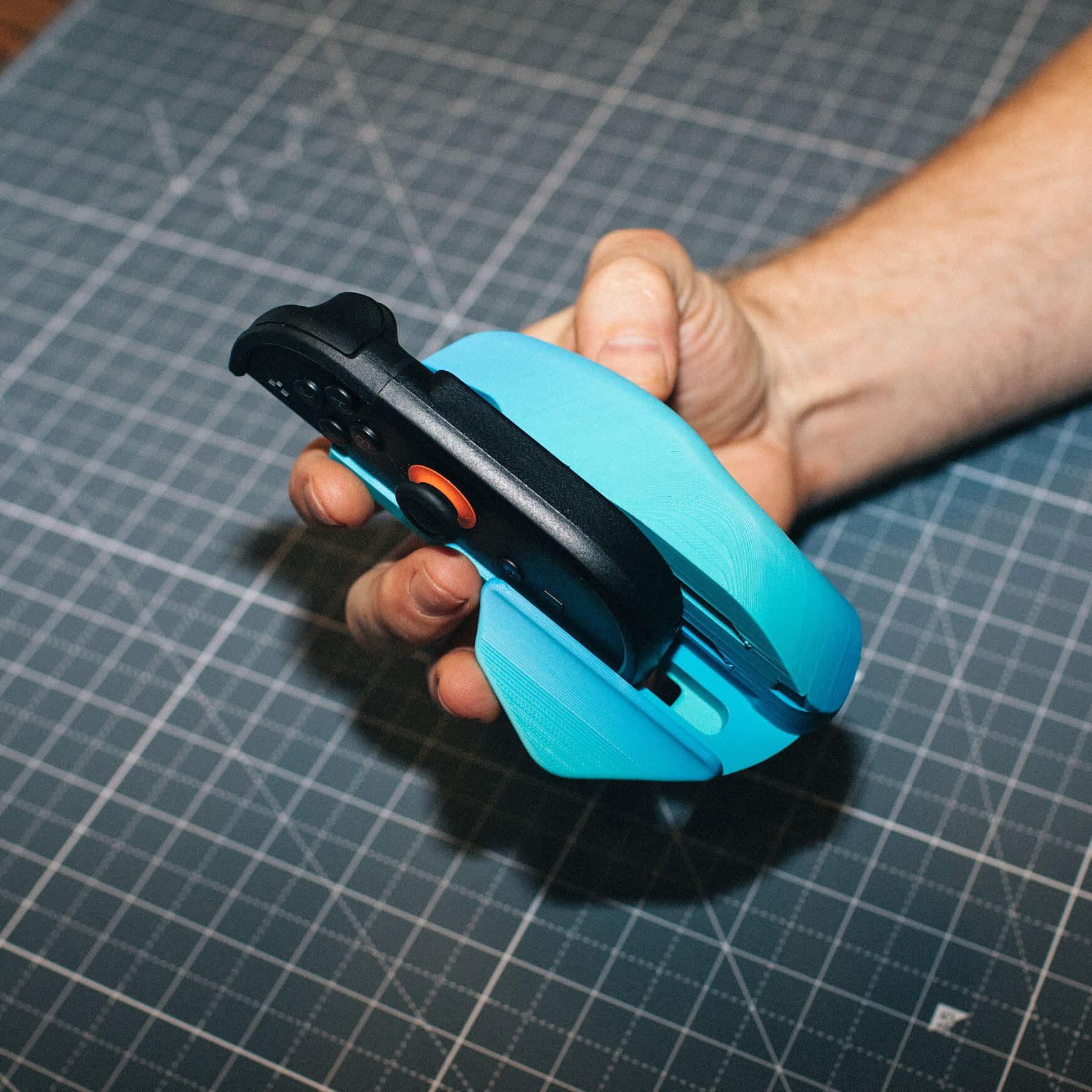 A hand holds a black gaming controller seated in a bright blue 3D-printed grip.