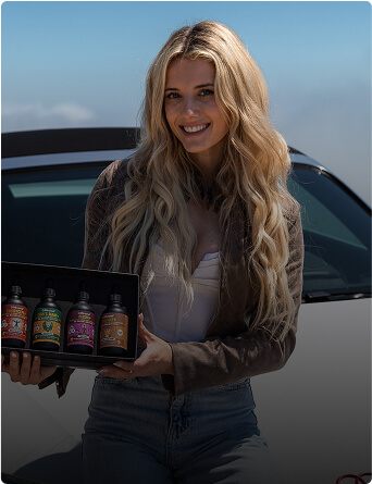 Person holding a box with four colorful bottles in front of a car.