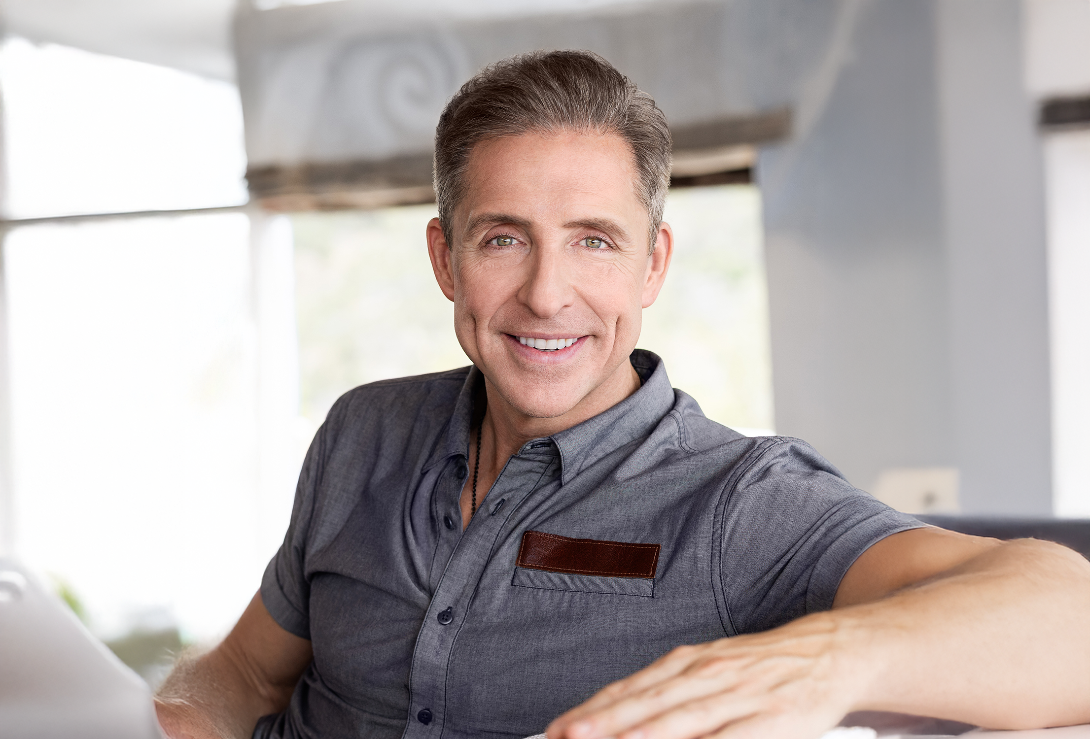 Smiling man in a gray shirt sitting indoors with natural light.