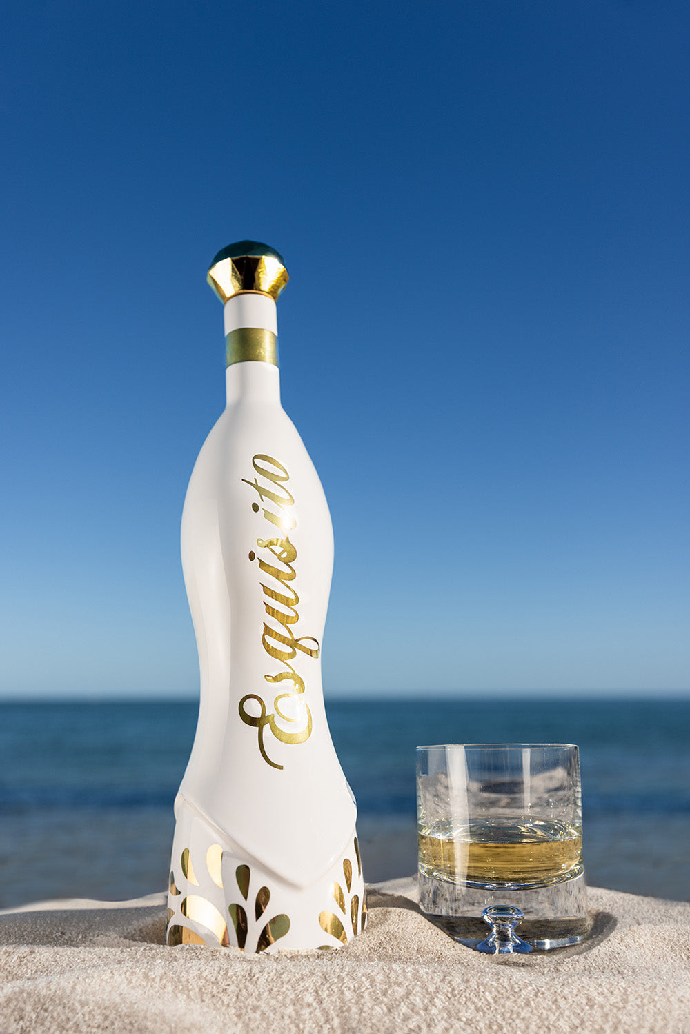 White bottle and glass on sandy beach with ocean background.