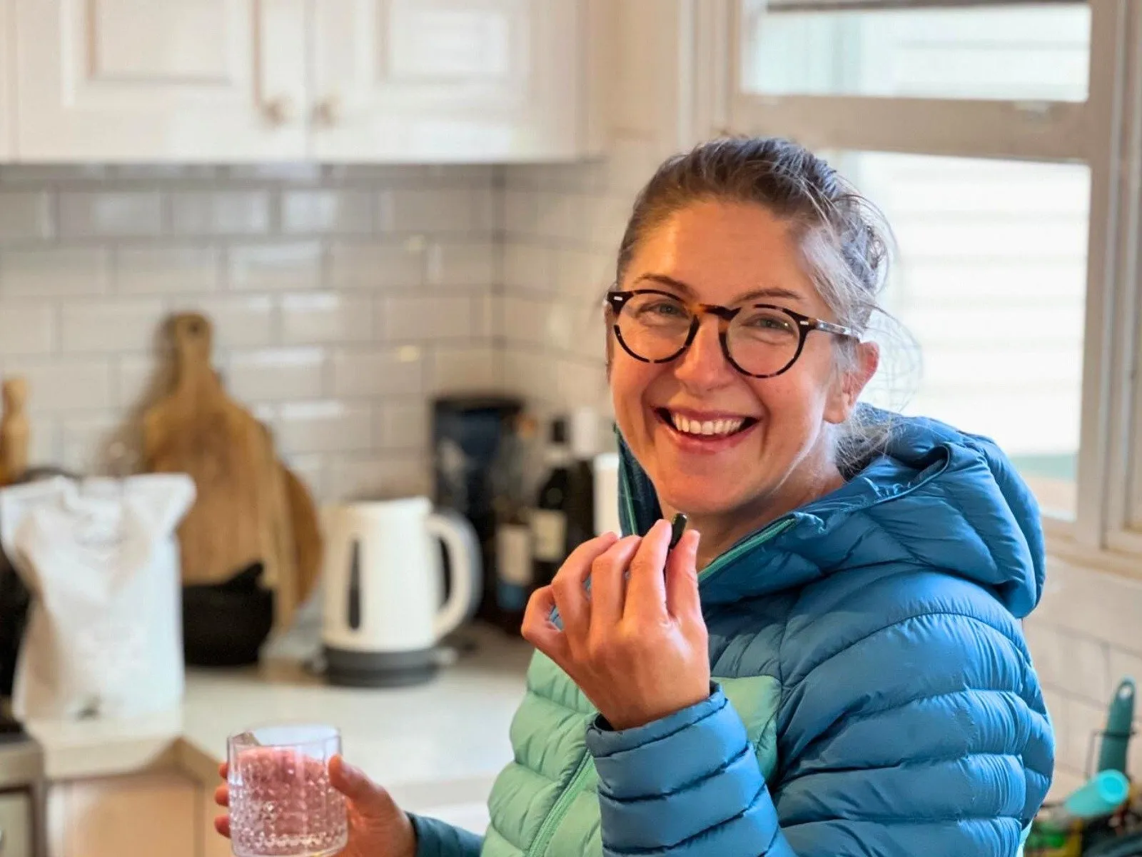 A smiling woman in a kitchen holds a dark capsule and a glass with a pink beverage.