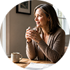 A woman sits at a table with a mug in her hands, looking out a window.