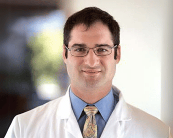 Man in glasses wearing a white coat and tie, smiling.