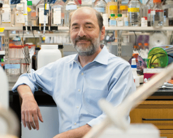 A person sitting in a laboratory with shelves of bottles and equipment in the background.