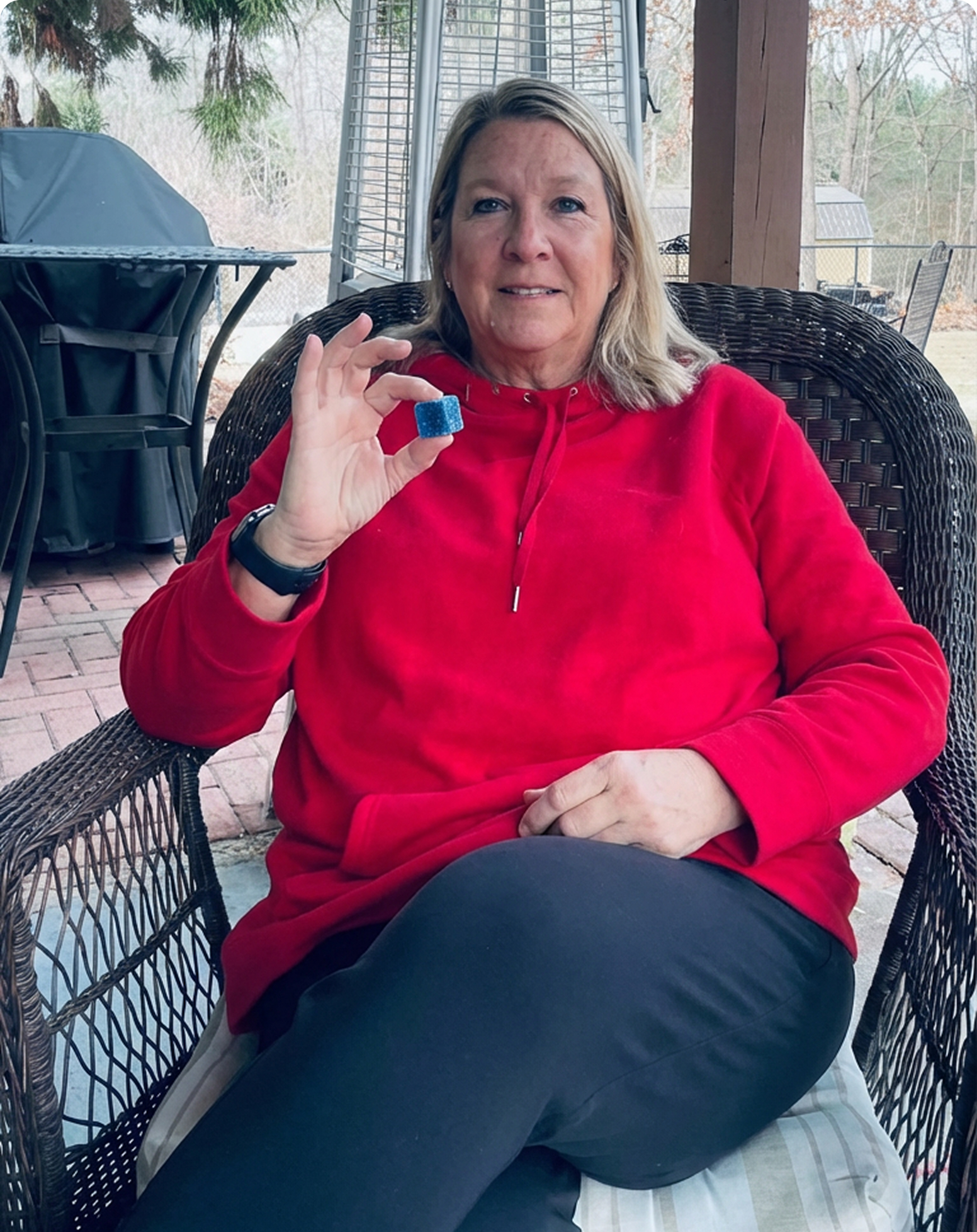 A woman in a red hoodie sits in a wicker chair, holding up a small, sparkly blue cube.