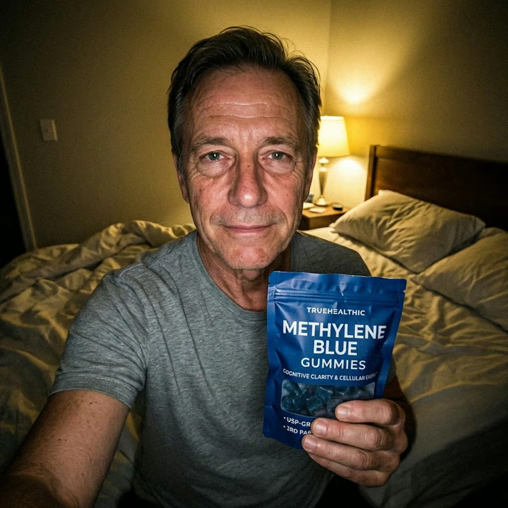 A man in a gray t-shirt sits on a bed holding up a blue pouch of Methylene Blue Gummies.