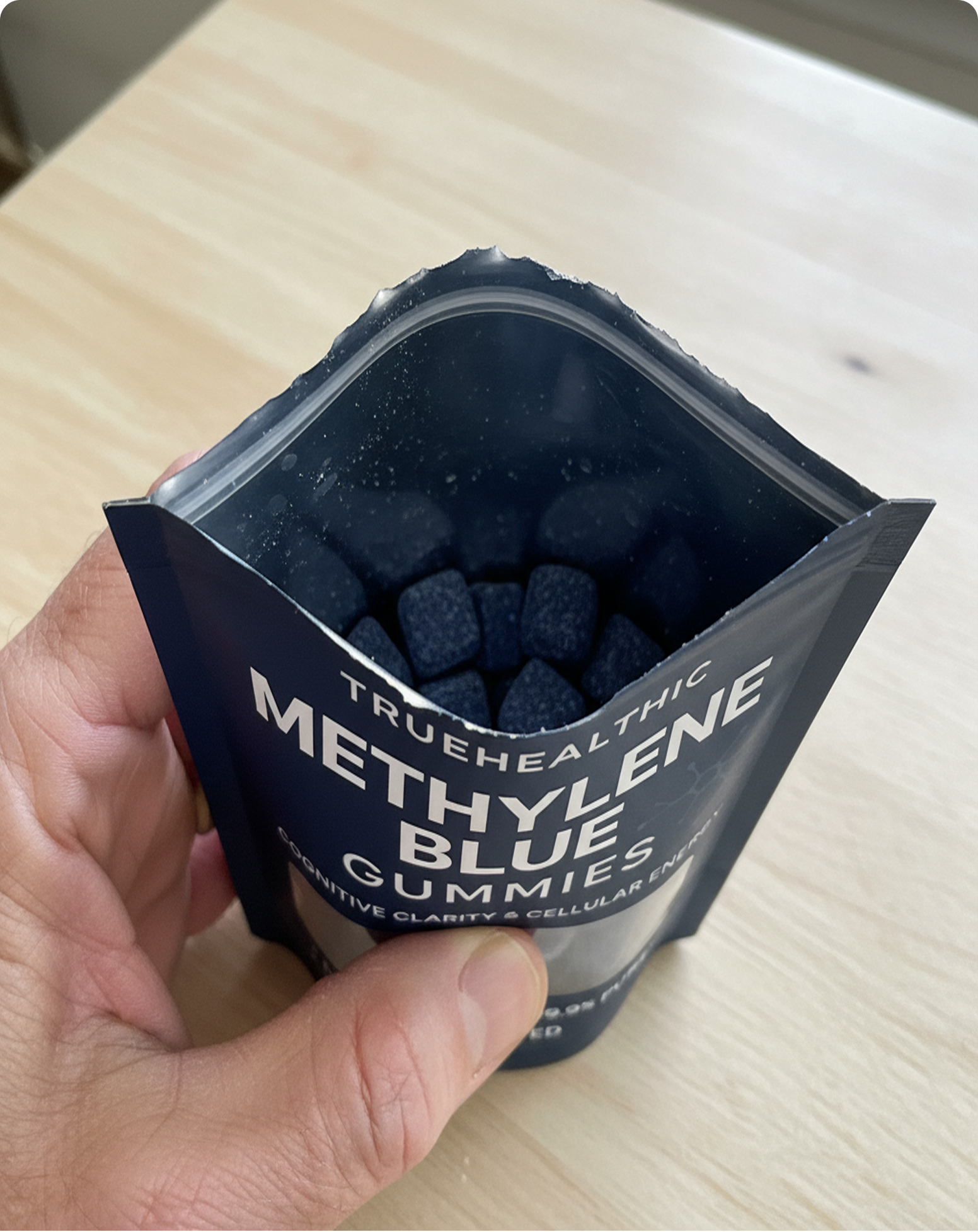 A hand holds open a dark blue pouch of Truehealthic Methylene Blue Gummies, revealing the contents.