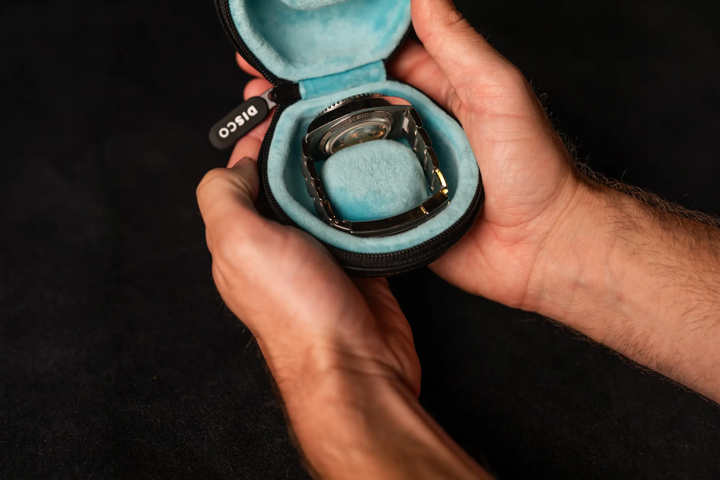 Two hands holding an open Black and Oslo Puck with a steel watch on its bracelet in the pale blue foam