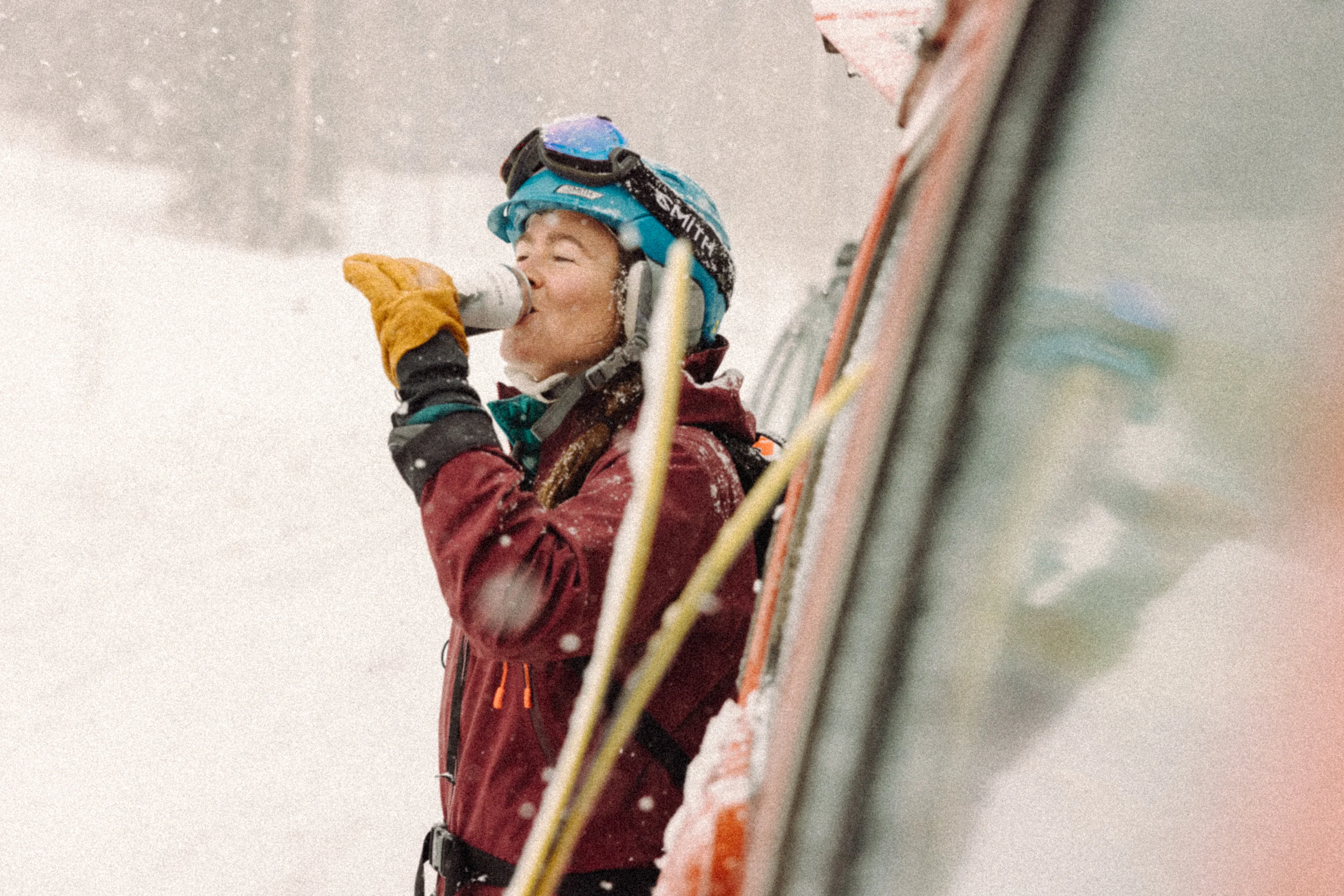 A person in a ski helmet and jacket drinks from a can in the snow.