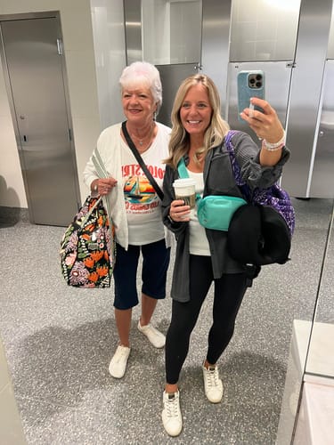 Two women smiling and posing for a mirror selfie in what appears to be a public restroom.
