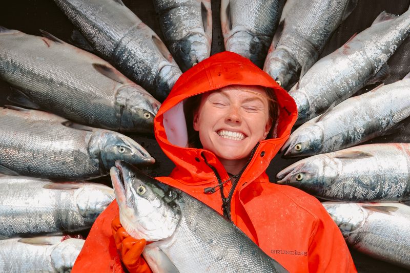Person in orange raincoat surrounded by fish, smiling with eyes closed.